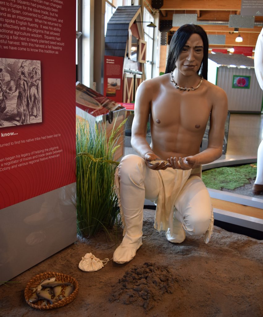 The Squanto mannequin in the recreated Plymouth Colony scene, wearing white leather trousers, a loincloth, and a necklace while holding a fish.