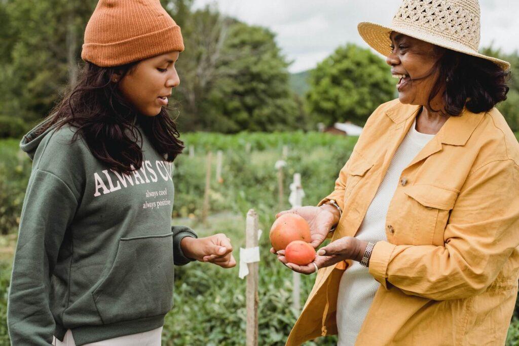 Female farmer smiling and showing vegetables to teenage girl asking a question