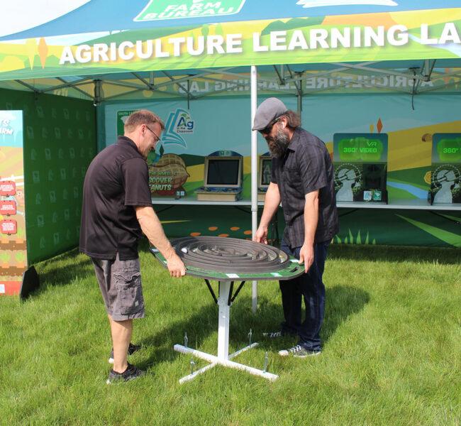 Two men playing the farm maze game in front of an exhibit tent