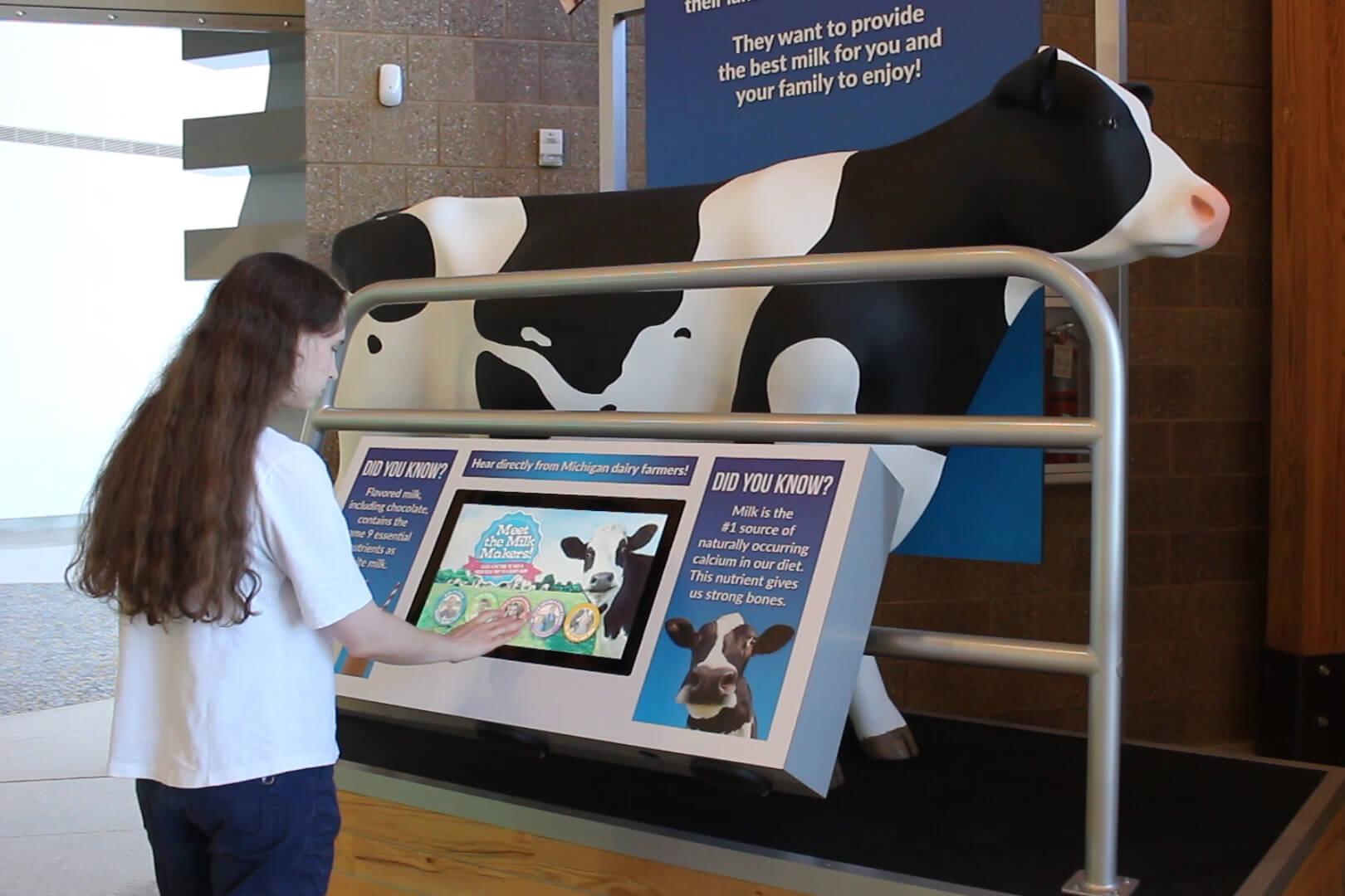 Woman standing near milking cow exhibit and interacting with video screen.