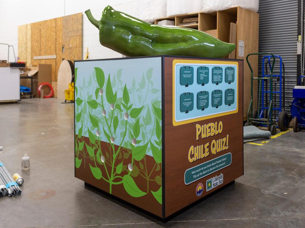 Chile pepper display, showing one side with trivia questions and one side with mini chile pepper replicas, with oversize pepper on top of display.
